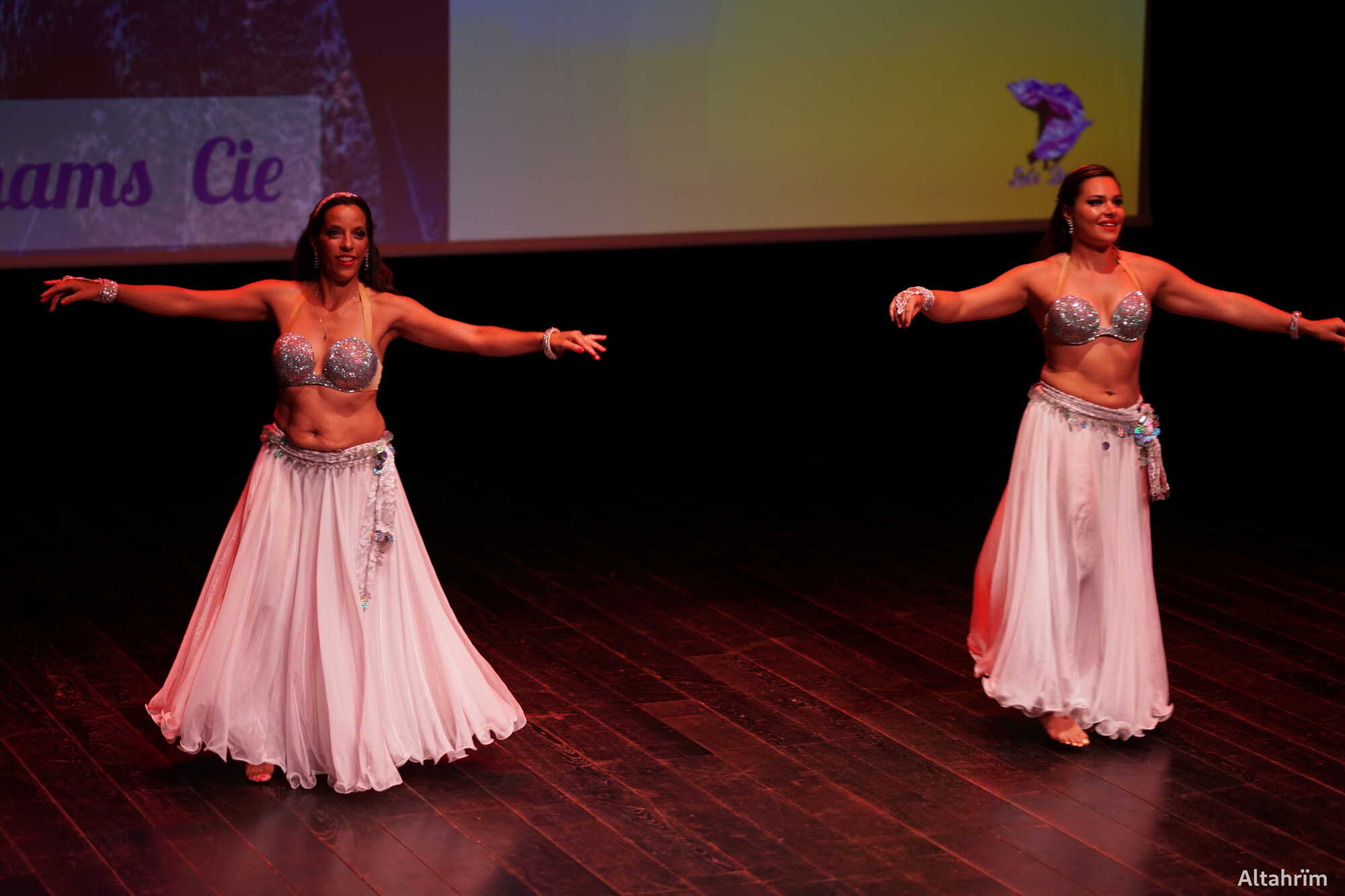 Deux femmes souriantes en costumes de danse orientale (soutien-gorge argenté, jupe blanche) sur scène, bras étendus. Écran avec "ams Cie".